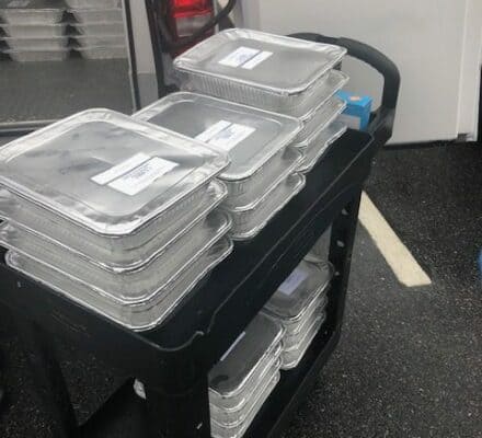 A cart stacked with covered aluminum food trays stands beside a vehicle, reminiscent of the organized displays in a luxury kitchen appliance showroom, with more trays visible inside the vehicle.
