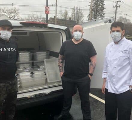 Three men wearing masks stand by an open vehicle trunk filled with plastic containers, perhaps delivering parts for a luxury kitchen appliance, on a cloudy day in a parking lot.