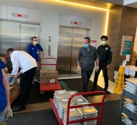 Several people, some wearing masks, unload food trays and boxes from carts in a building lobby near elevators, possibly delivering to a kitchen showroom featuring Wolf and Sub-Zero appliances.