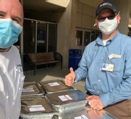 Two men wearing masks and work attire stand outdoors by trays of food containers, giving a thumbs up, perhaps after a meal delivery near a kitchen showroom.
