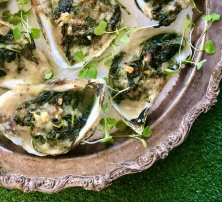 A silver platter holds baked oysters topped with herbs and microgreens, artfully arranged as if served fresh from a luxury kitchen appliance, on a green textured surface.