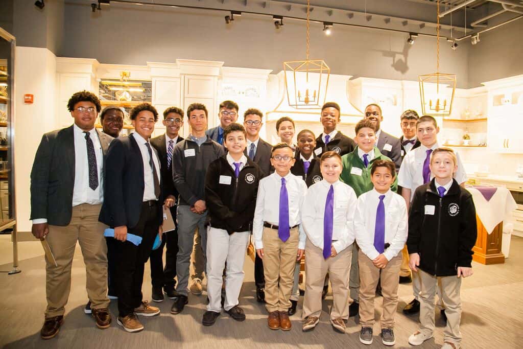 A group of boys and teenagers, dressed in semi-formal attire with ties and jackets, pose for a group photo indoors under bright lighting in a modern appliance showroom.