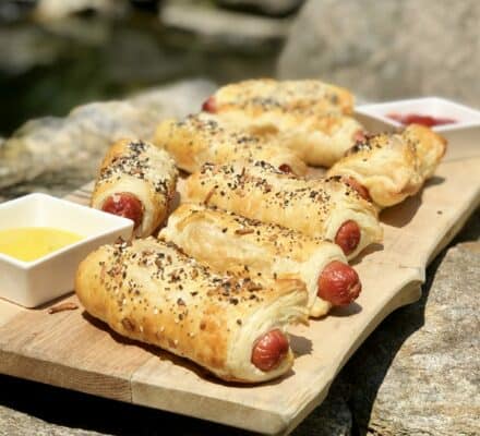 Six puff pastry-wrapped hot dogs with seasoning are arranged on a wooden board, alongside two small bowls of mustard and ketchup, set on a stone surface outdoors—perfect inspiration for your next kitchen showroom or luxury kitchen appliance demo.