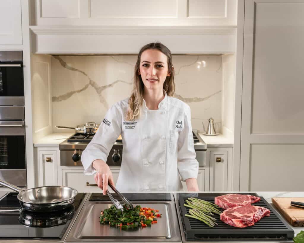 A chef in a white uniform uses tongs to cook vegetables on a stovetop, with asparagus and raw steaks beside her in a modern kitchen showroom featuring sleek Sub Zero appliances.