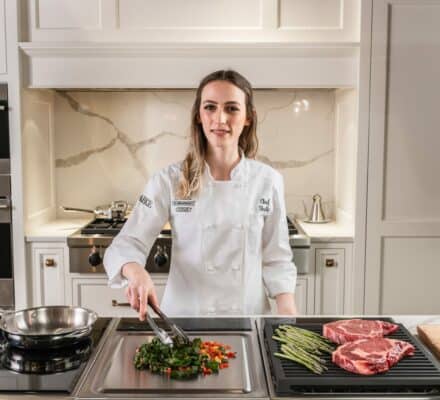 A chef in a white uniform uses tongs to cook vegetables on a stovetop, with asparagus and raw steaks beside her in a modern kitchen showroom featuring sleek Sub Zero appliances.