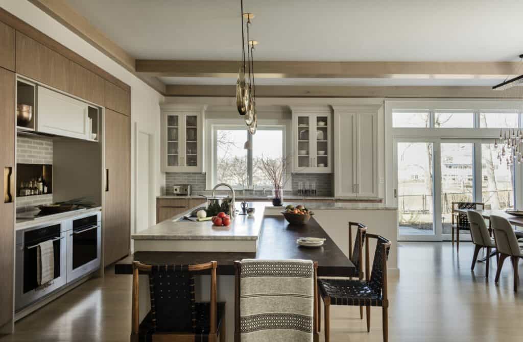 Modern kitchen showroom featuring wood and white cabinetry, a large island with barstools, pendant lights, built-in Sub Zero and Wolf appliances, plus floor-to-ceiling windows pouring in natural light.