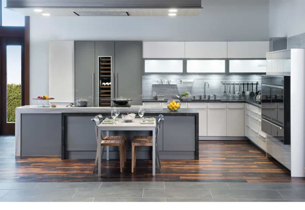 Modern kitchen showroom with gray and white cabinets, luxury kitchen appliances, a central island with cooktop, and a dining table set for two beneath bright overhead lighting.