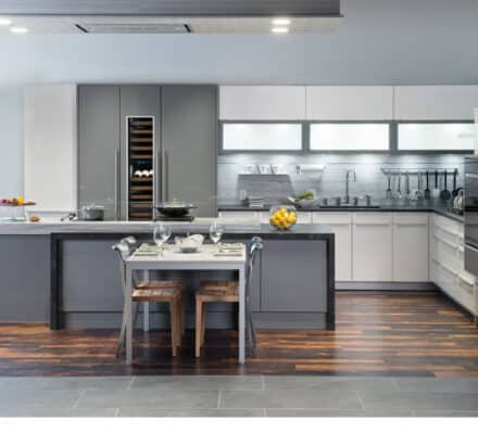 Modern kitchen showroom with gray and white cabinets, luxury kitchen appliances, a central island with cooktop, and a dining table set for two beneath bright overhead lighting.