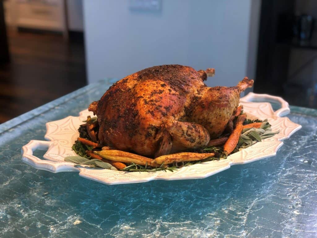 A whole roasted chicken seasoned with herbs is served on a white platter with carrots and greens, elegantly displayed as you’d find in a kitchen showroom featuring a Wolf appliance.