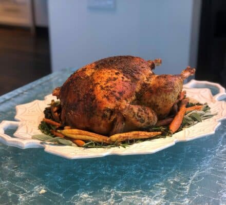 A whole roasted chicken seasoned with herbs is served on a white platter with carrots and greens, elegantly displayed as you’d find in a kitchen showroom featuring a Wolf appliance.