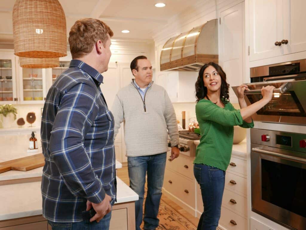 Three adults stand in a kitchen; one woman opens an oven while two men watch her. The kitchen features light cabinets, modern design, and a luxury kitchen appliance that adds sophistication to the scene.