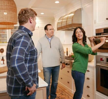 Three adults stand in a kitchen; one woman opens an oven while two men watch her. The kitchen features light cabinets, modern design, and a luxury kitchen appliance that adds sophistication to the scene.