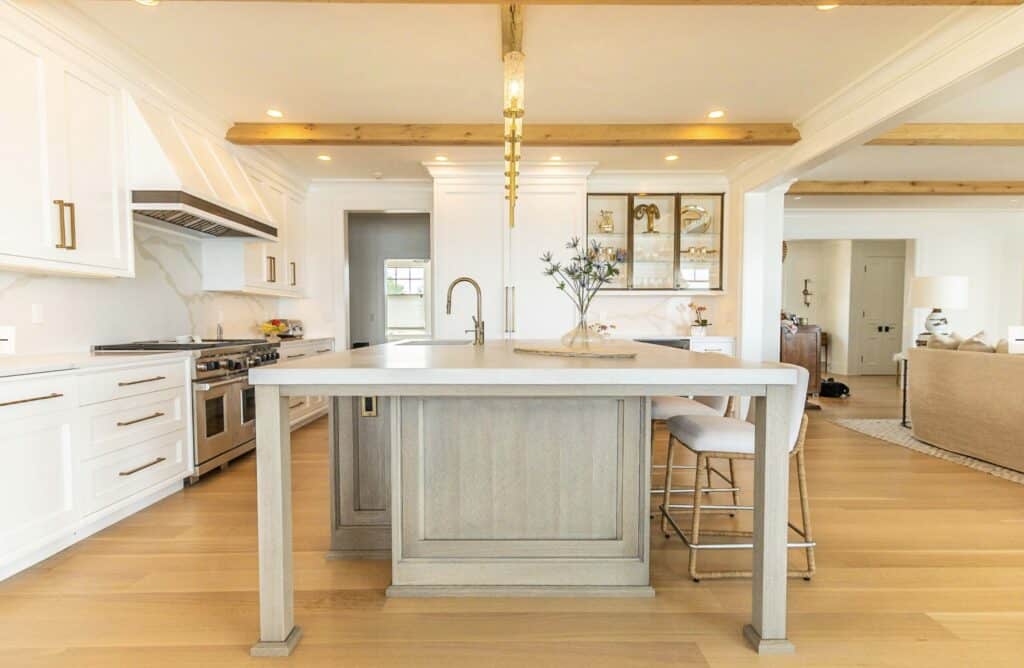 Bright, modern kitchen with white cabinets, a luxury kitchen appliance suite including Wolf appliances, a light wood island, two upholstered stools, and wood beams on the ceiling. Living area visible in the background.