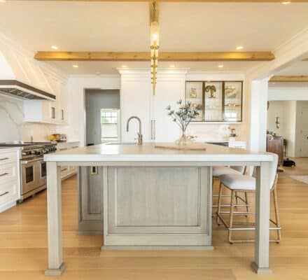 Bright, modern kitchen with white cabinets, a luxury kitchen appliance suite including Wolf appliances, a light wood island, two upholstered stools, and wood beams on the ceiling. Living area visible in the background.