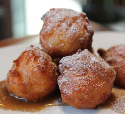 Four golden-brown fritters dusted with powdered sugar and drizzled with syrup are artfully arranged on a white plate—perfectly prepared using a luxury kitchen appliance.