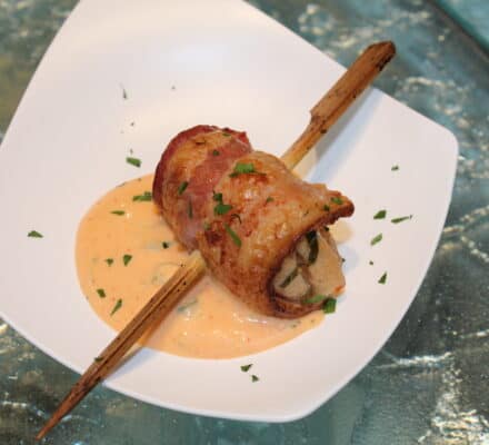 A bacon-wrapped roll stuffed with vegetables is skewered and served on a white plate with a creamy orange sauce, garnished with chopped herbs—perfectly prepared using a luxury kitchen appliance.