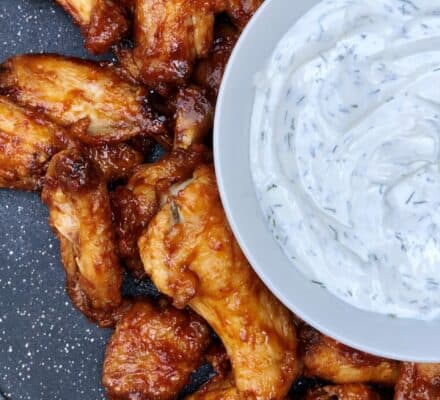 Barbecue chicken wings arranged on a dark surface next to a bowl of creamy white dipping sauce with herbs, perfect for serving in a luxury kitchen appliance demo or at your next gathering inspired by an upscale appliance showroom.