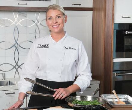 A chef in a white uniform stands in a modern appliance showroom kitchen holding tongs, with cooked green vegetables in a pan on the counter.