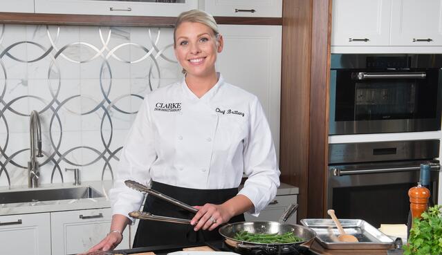 A chef in a white uniform stands in a modern appliance showroom kitchen holding tongs, with cooked green vegetables in a pan on the counter.