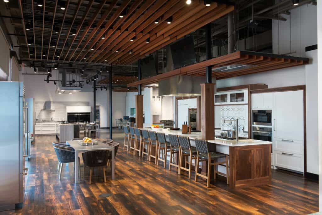Modern, spacious kitchen with wooden flooring, a large island with bar stools, a dining table, Sub Zero appliance, stainless steel finishes, and an open ceiling with exposed beams and lighting—perfect for a kitchen showroom.