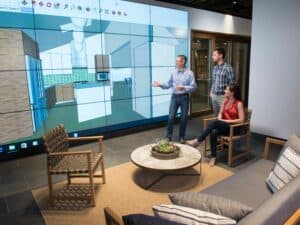 Three people review a 3D kitchen design, featuring luxury kitchen appliances, displayed on a large screen in a modern office setting with lounge furniture.