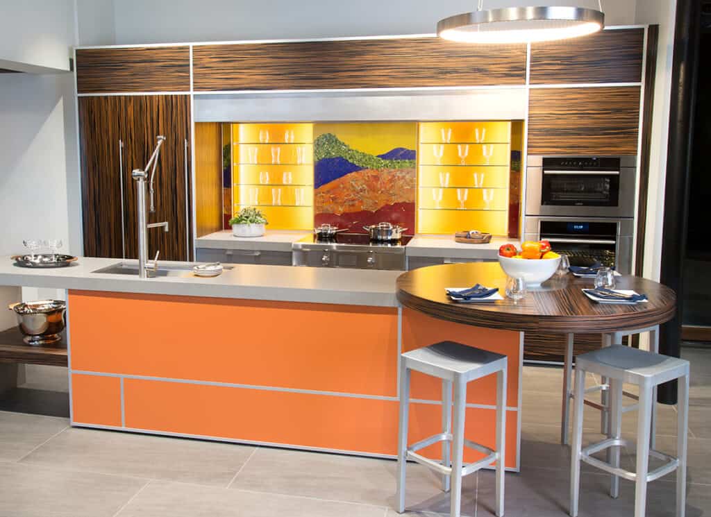 Modern kitchen with orange and wood accents, Sub-Zero and Wolf appliances, a round dining area, and a colorful backsplash depicting a landscape—perfect inspiration for any kitchen showroom.