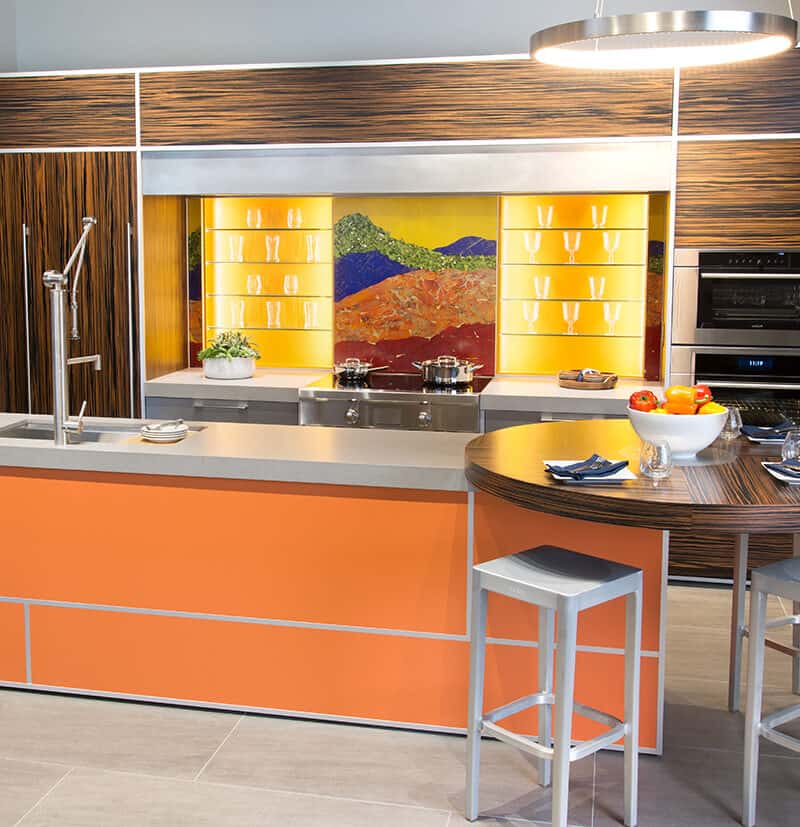 Modern kitchen with orange and wood accents, Sub-Zero and Wolf appliances, a round dining area, and a colorful backsplash depicting a landscape—perfect inspiration for any kitchen showroom.