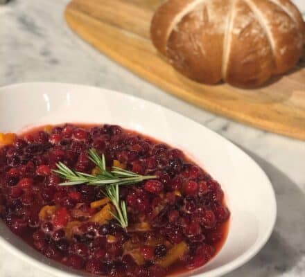 A white bowl of cranberry sauce garnished with rosemary sits on a marble counter, highlighting the elegance of a kitchen featuring Sub Zero appliances, with a round loaf of bread on a wooden board in the background.