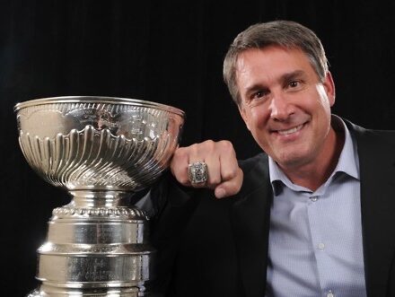 A man in a suit smiles beside a large silver trophy, proudly displaying a ring on his finger, as if celebrating his success in an upscale appliance showroom specializing in luxury kitchen appliances.