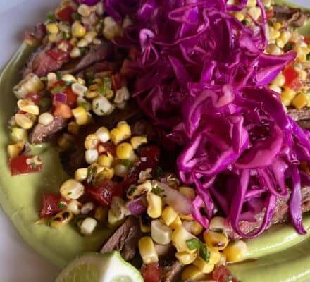 A dish featuring sliced steak on a bed of creamy green sauce, topped with corn salsa and shredded purple cabbage, garnished with lime wedges—perfect inspiration for your next visit to a luxury kitchen appliance showroom.