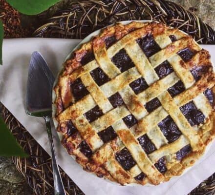 A baked pie with a golden-brown lattice crust sits on a wicker tray next to a metal pie server, atop a folded white napkin—ready to serve from your luxury kitchen appliance.