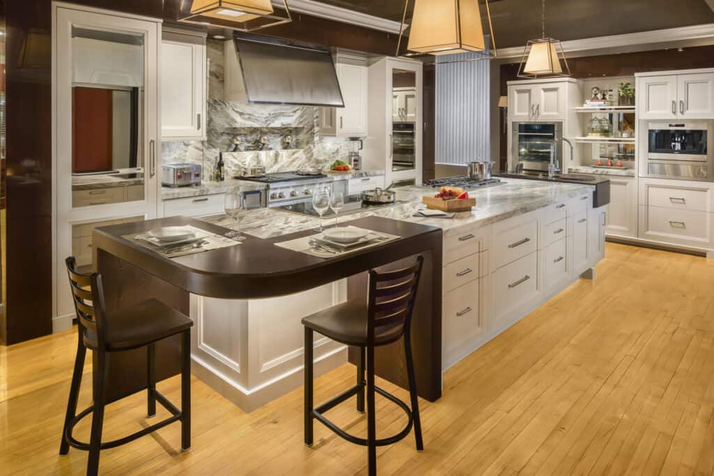 Modern kitchen with white cabinets, marble countertops, stainless steel appliances—including a Wolf appliance—island seating for two, and pendant lighting on a light wood floor.