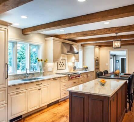 A spacious kitchen showroom with white cabinets, wooden beams, an island with seating, stainless steel appliances—including a premium Wolf appliance—and a stone fireplace in the background.