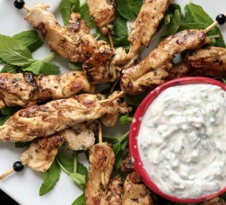 Grilled chicken skewers arranged on fresh mint leaves next to a bowl of creamy herb dip, served on a white square plate—perfect inspiration for your next gathering from an appliance showroom featuring Wolf and Sub Zero appliances.