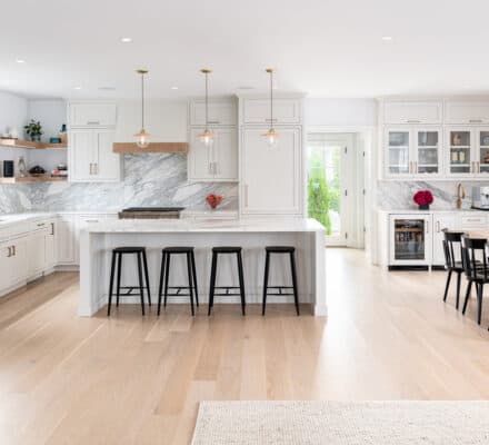 Spacious modern kitchen showroom with white cabinets, marble countertops, a large island with four black stools, and a wooden dining table with six black chairs. Ample natural light and luxury kitchen appliance upgrades throughout.