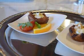 A metal tray holds a white plate with a stuffed mushroom, a slice of yellow bell pepper, a cherry tomato half, and a sprig of parsley—perfect for tasting at your local kitchen showroom.