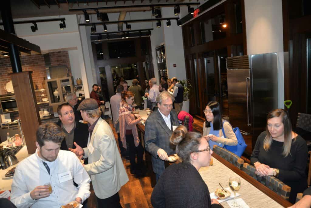 A group of people socialize and eat at a professional indoor event, standing and sitting around a kitchen island and communal tables in a stunning kitchen showroom featuring luxury kitchen appliances.