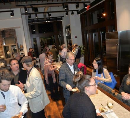 A group of people socialize and eat at a professional indoor event, standing and sitting around a kitchen island and communal tables in a stunning kitchen showroom featuring luxury kitchen appliances.