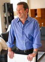 A man in a blue button-up shirt stands indoors, smiling, with hands resting on a surface. Modern furniture and a luxury kitchen appliance, including a Sub-Zero appliance, are visible in the background.