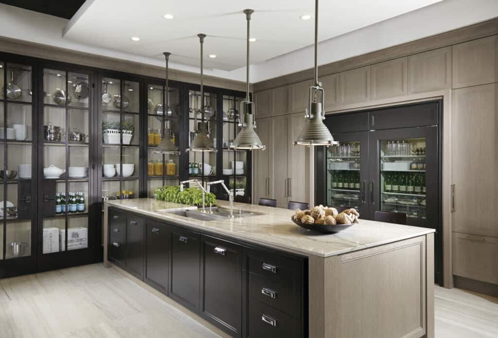 Modern kitchen with a large island, pendant lights, glass-front cabinets displaying dishes and glassware, and luxury kitchen appliances including double-door refrigerators in the background.