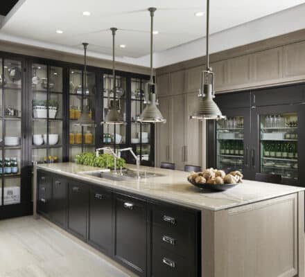 Modern kitchen with a large island, pendant lights, glass-front cabinets displaying dishes and glassware, and luxury kitchen appliances including double-door refrigerators in the background.