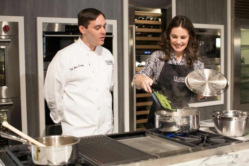 A woman adds green beans to a pot on a luxury kitchen appliance while a man in a chef's coat watches in a modern kitchen setting.