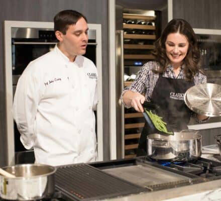 A woman adds green beans to a pot on a luxury kitchen appliance while a man in a chef's coat watches in a modern kitchen setting.