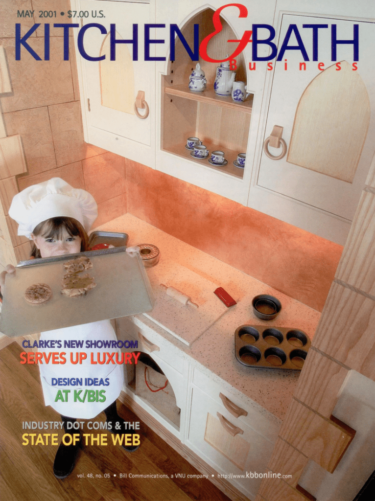 A young girl in a chef hat holds a baking tray in a modern kitchen with a luxury kitchen appliance, featured on the May 2001 cover of Kitchen & Bath Business magazine.