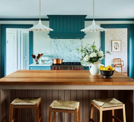 A kitchen with a wooden island, three stools, blue cabinetry, marble backsplash, pendant lights, and a bowl of lemons next to a white pitcher with flowers features luxury kitchen appliances for a refined touch.