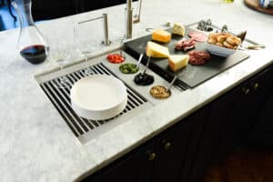 Charcuterie board with assorted cheeses, meats, crackers, and olives sits on a marble countertop in an appliance showroom, next to stacked plates, wine glasses, and a half-filled wine decanter.