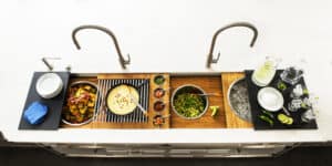 Overhead view of a modern kitchen sink with two faucets and prepared food, drinks, bowls, and ingredients arranged neatly on the countertop in a stylish kitchen showroom featuring Sub-Zero and Wolf appliances.