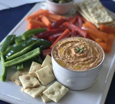 A platter with a bowl of hummus, surrounded by green peppers, red peppers, orange peppers, carrots, crackers, and a dish of green olives in the background—perfect for serving guests in a kitchen equipped with a luxury Wolf appliance.
