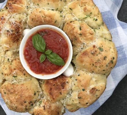 A ring of pull-apart garlic bread with grated cheese and herbs surrounds a bowl of marinara sauce garnished with basil, set on a blue and white cloth in a stunning kitchen showroom featuring Wolf and Sub-Zero appliances.