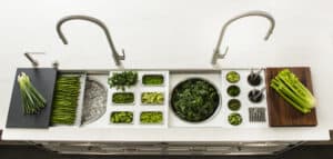 Top-down view of a modern kitchen sink with metal faucets, various green vegetables, and herbs arranged neatly in trays and bowls along the counter, evoking the look of a luxury kitchen appliance showroom.